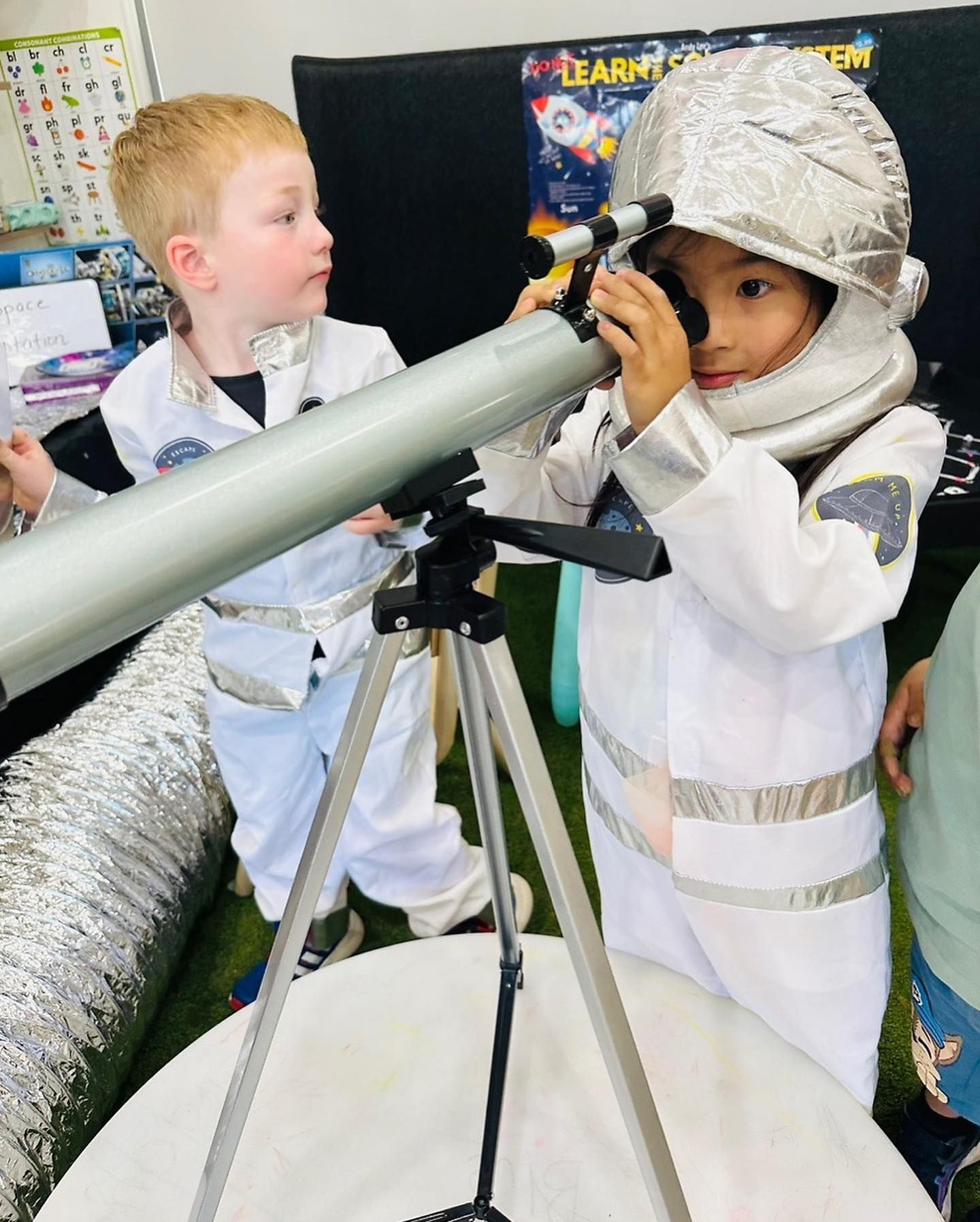 Two children in astronaut costumes look through a telescope in a classroom setting with space-themed decorations.