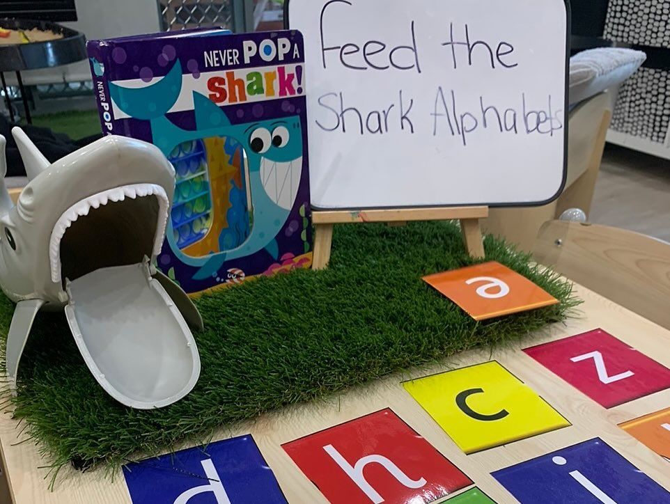 A toy shark with an open mouth sits on artificial grass beside alphabet cards and a sign reading "Feed the Shark Alphabets."