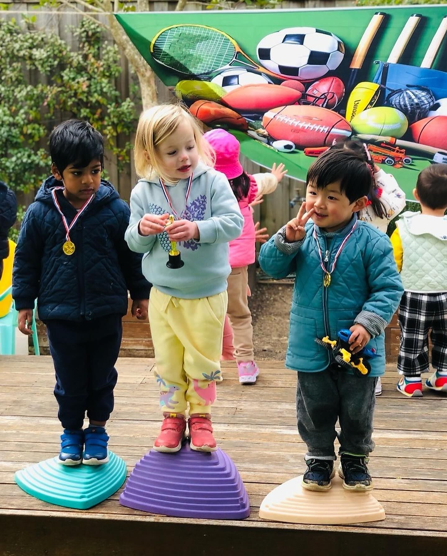 Three children stand on individual plastic platforms on a stage, posing with medals and trophies in front of sports gear.