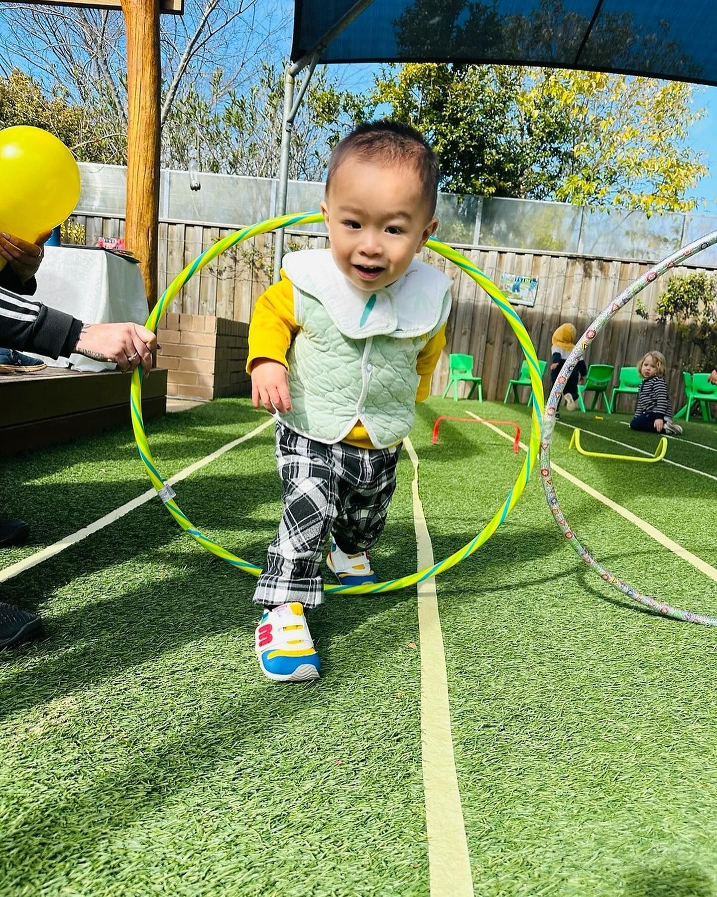 A smiling child walking through a colorful hula hoop on a green outdoor play area with other children in the background.