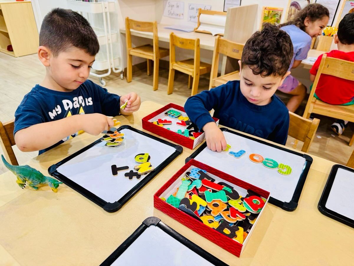 Children sit at a table in a classroom, placing colorful magnetic letters onto whiteboards.