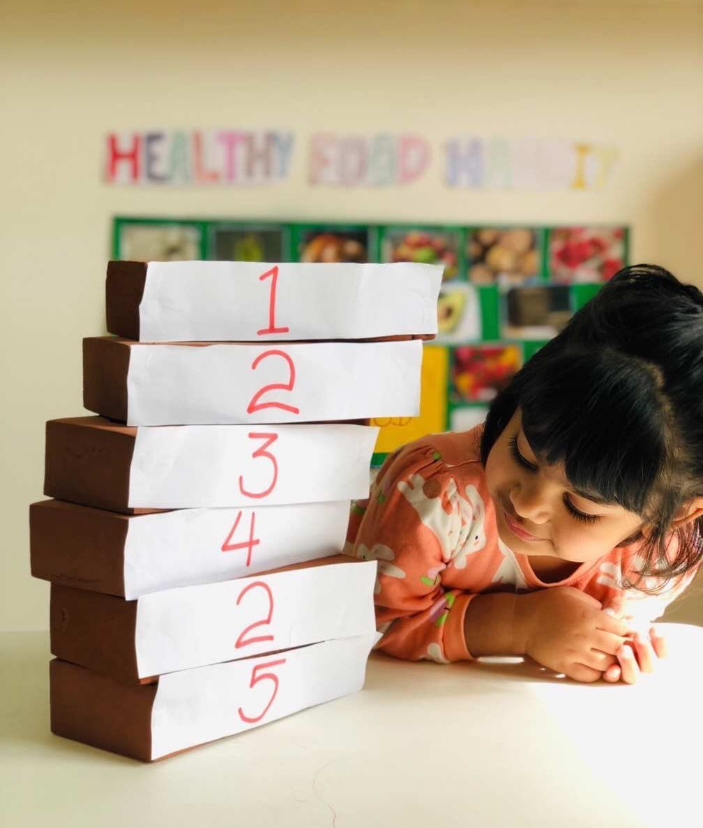 A child observes a vertical stack of six brown blocks, each labeled with a hand-written number, in a classroom setting.