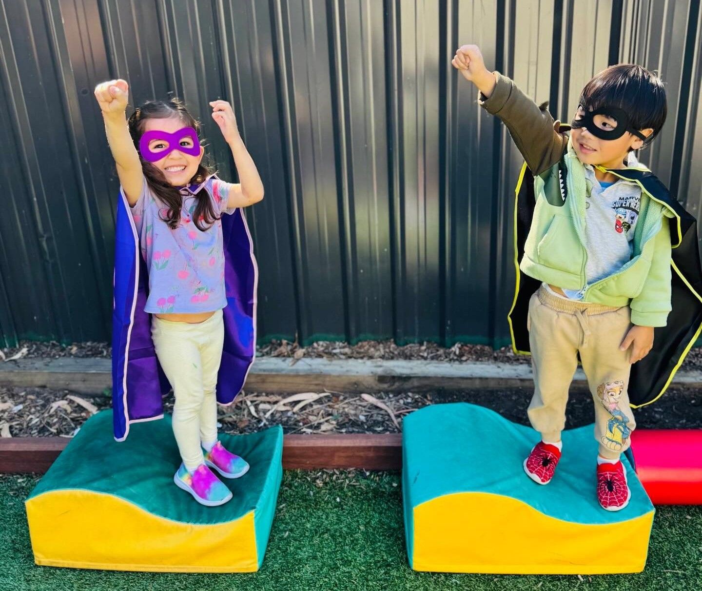 Two children in costumes standing on colorful platforms, raising their arms in front of a dark fence.