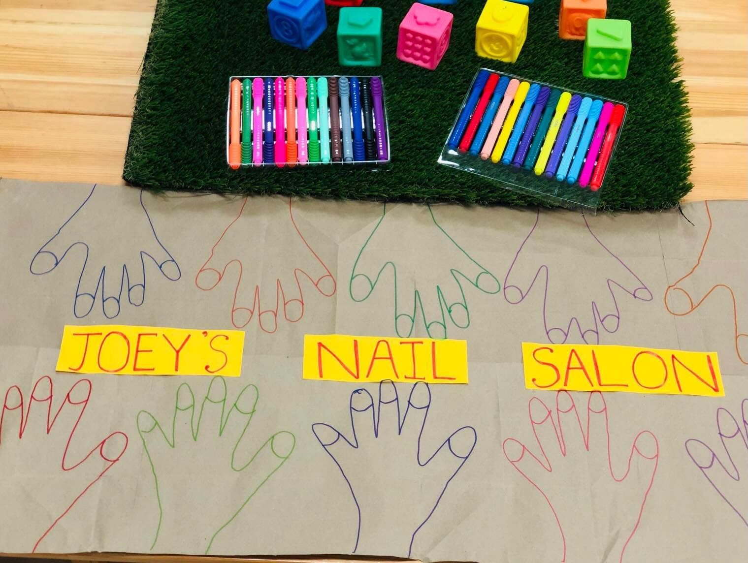 Children’s hand outlines and labeled blocks on paper beside colorful markers and toys on a mat