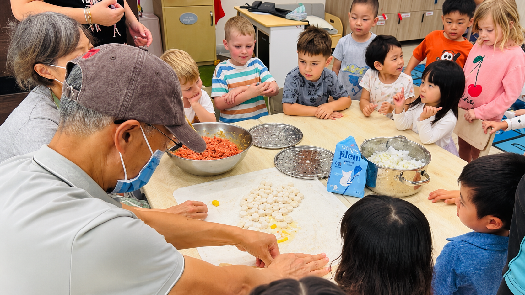 Three people in masks knead dough at a table in a bright kitchen classroom.