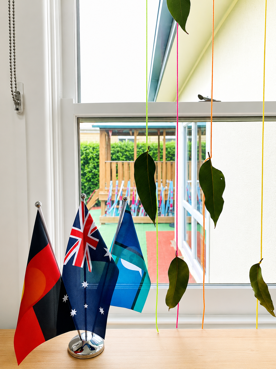 Small flags on a windowsill with hanging leaves, looking out onto a colorful playground.