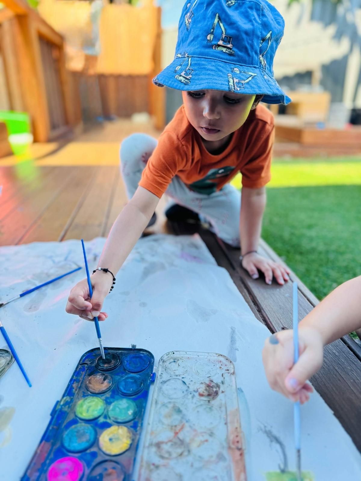 A child in a blue bucket hat paints on large paper outdoors using a watercolor set.