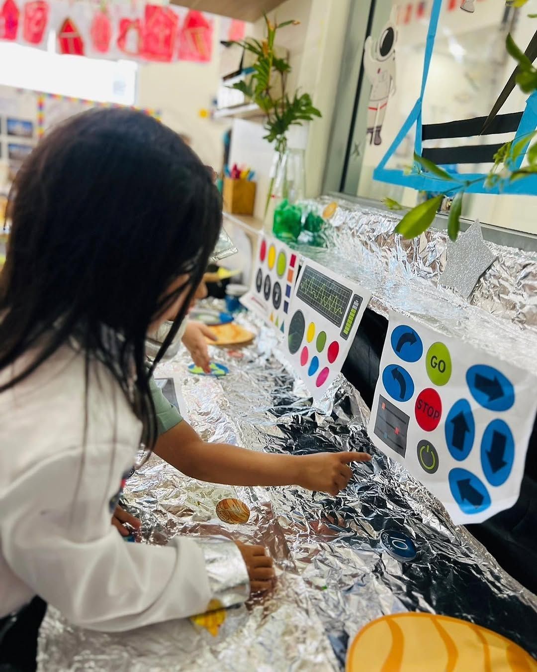 A child in a lab coat interacts with a foil-covered console featuring colorful, labeled buttons and directional arrows.