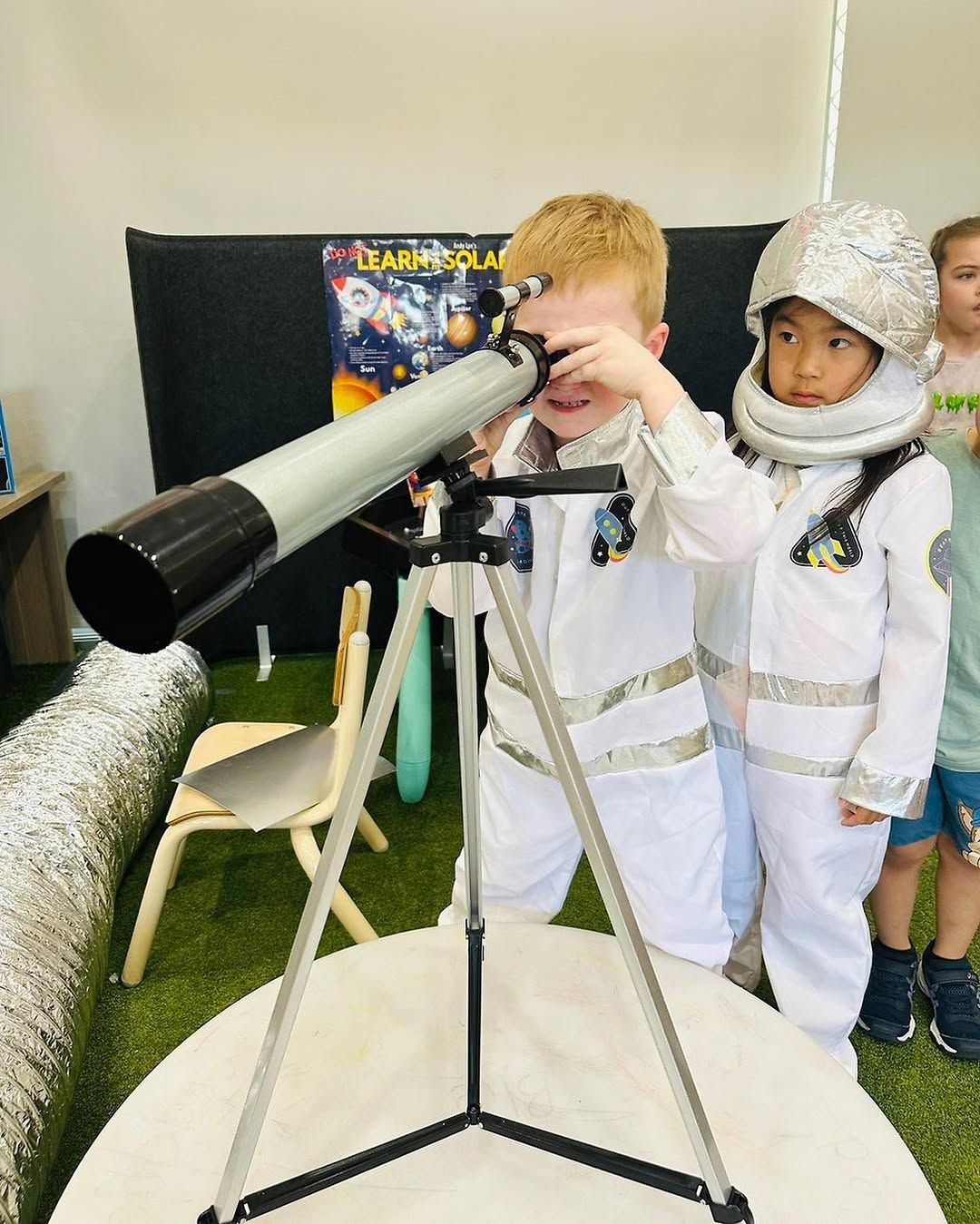Two people in white astronaut costumes look through a telescope on a tripod in a room decorated with space-themed posters.
