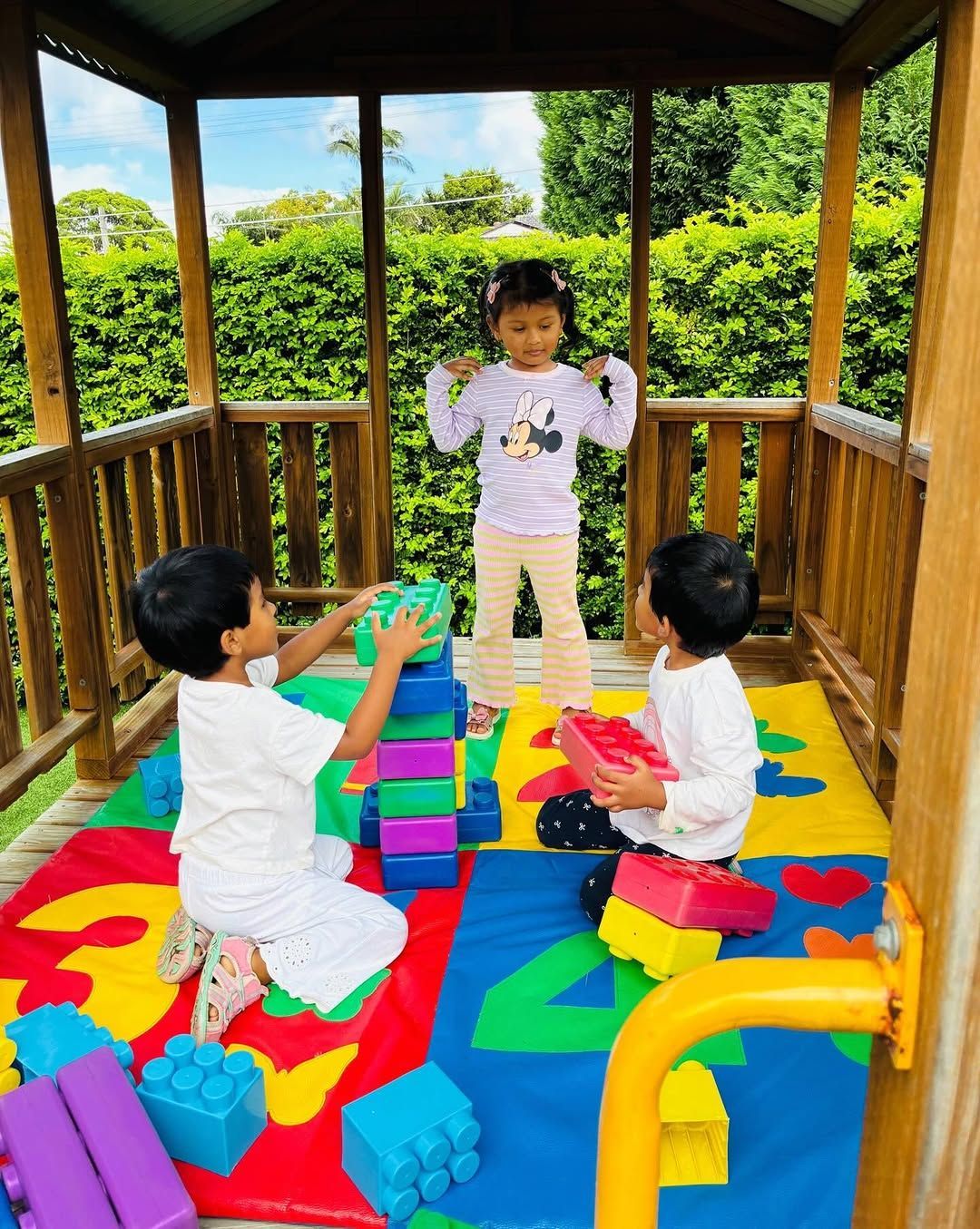 Three children play with colorful plastic building blocks inside a wooden, open-air playhouse with a mat on the floor.