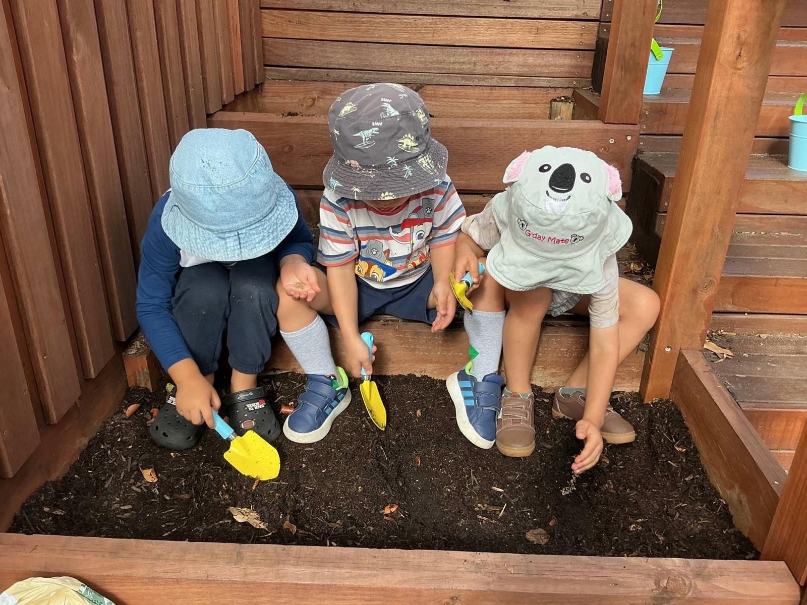 Three children wearing sun hats sit on a wooden step, digging in a soil box with colorful plastic shovels.