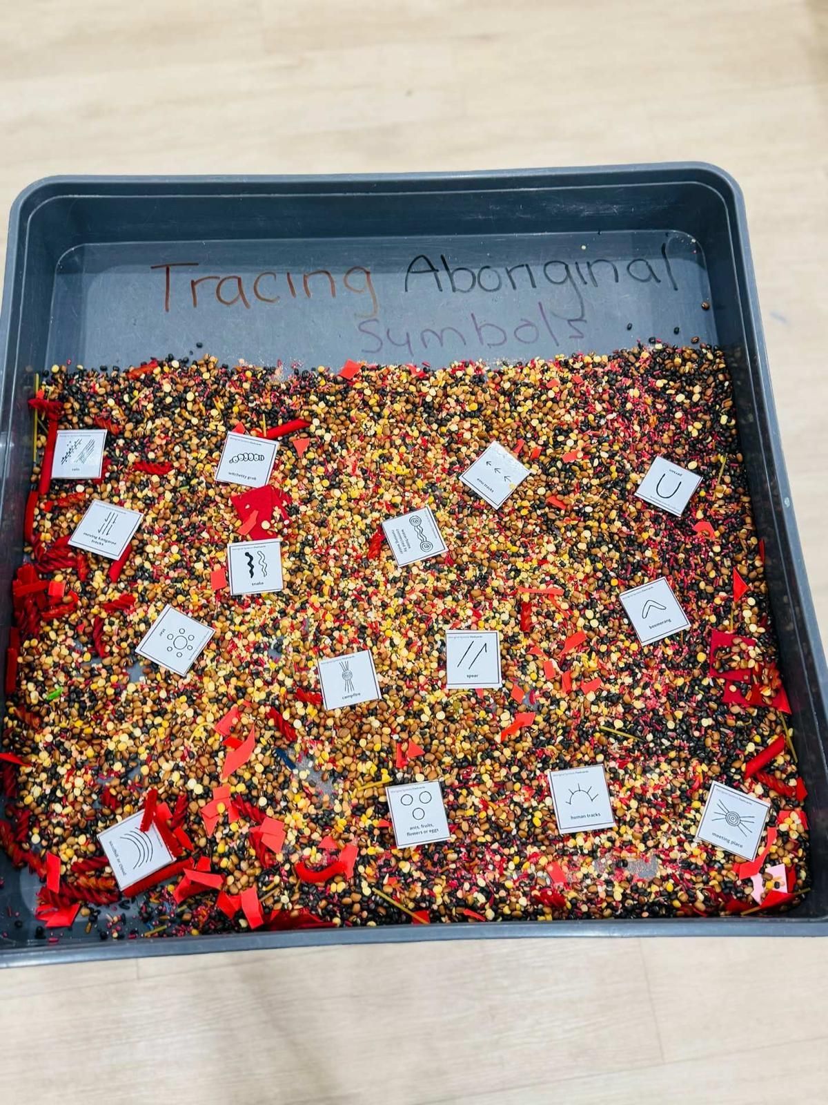 A dark tray filled with colorful grain and small cards featuring Aboriginal symbols for an educational tracing activity.