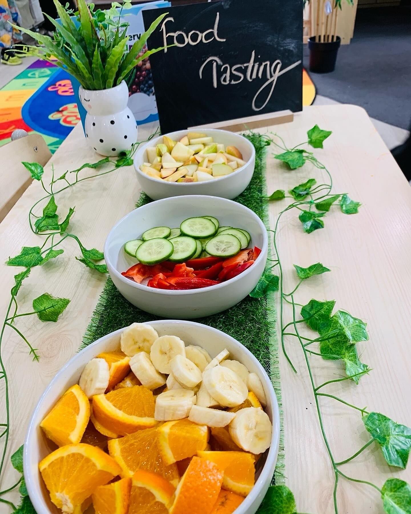 Three white bowls filled with sliced fresh fruit and vegetables are arranged on a table for a food tasting event.
