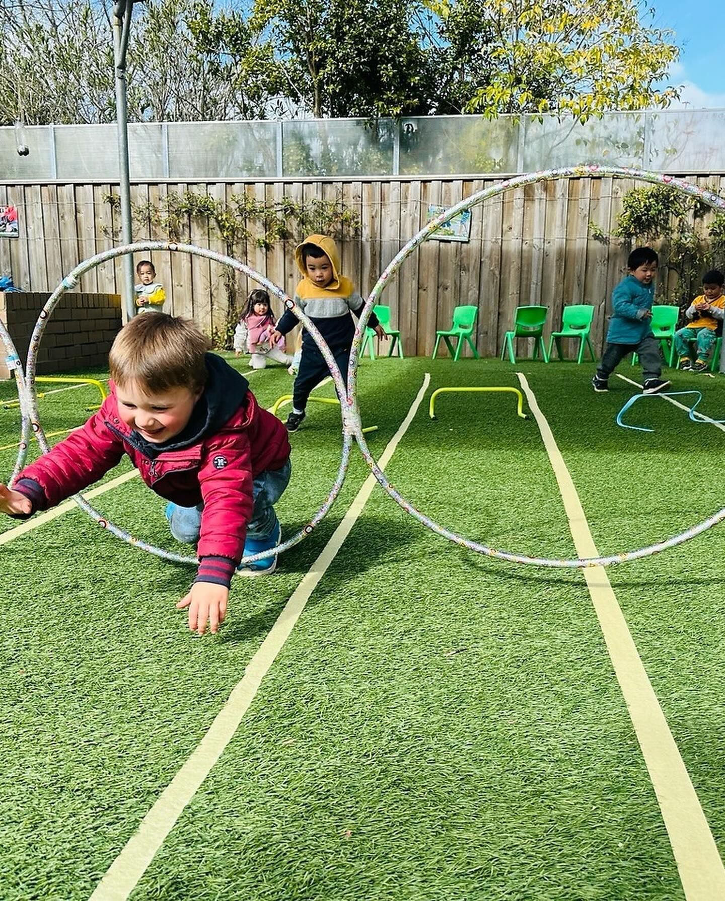 Children play outdoors on an artificial turf field with hoops and exercise equipment on a sunny day.