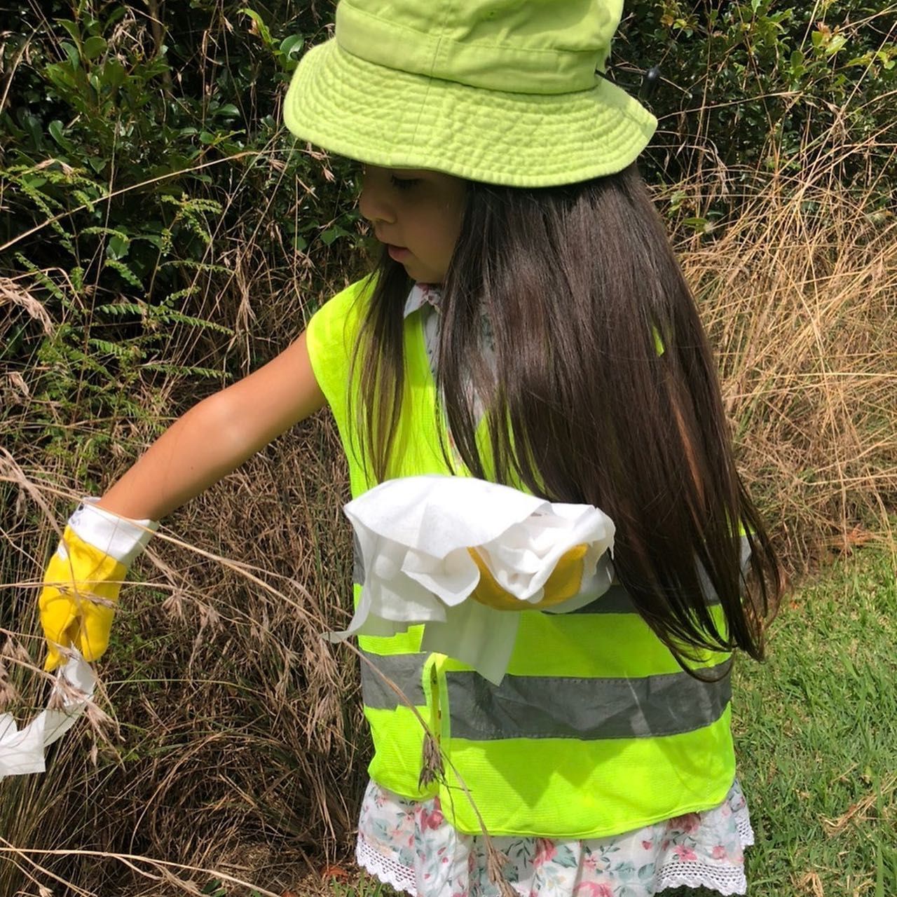 A person wearing a neon yellow safety vest, hat, and gloves picks up trash in a grassy, brush-filled area.