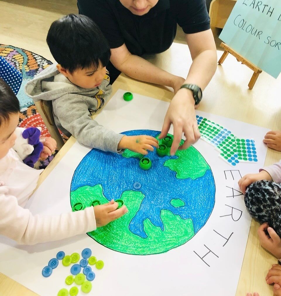 Children and an adult place blue and green bottle caps on a drawn Earth activity sheet labeled "Earth Day Color Sort."