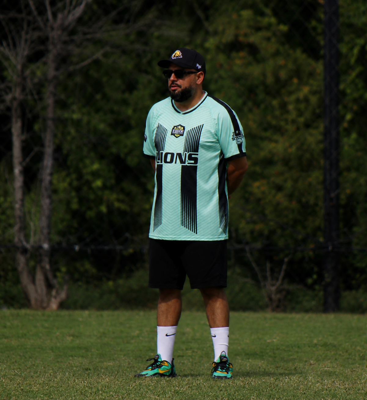 Man standing on grass in a light blue sports jersey and black shorts, facing left outdoors.