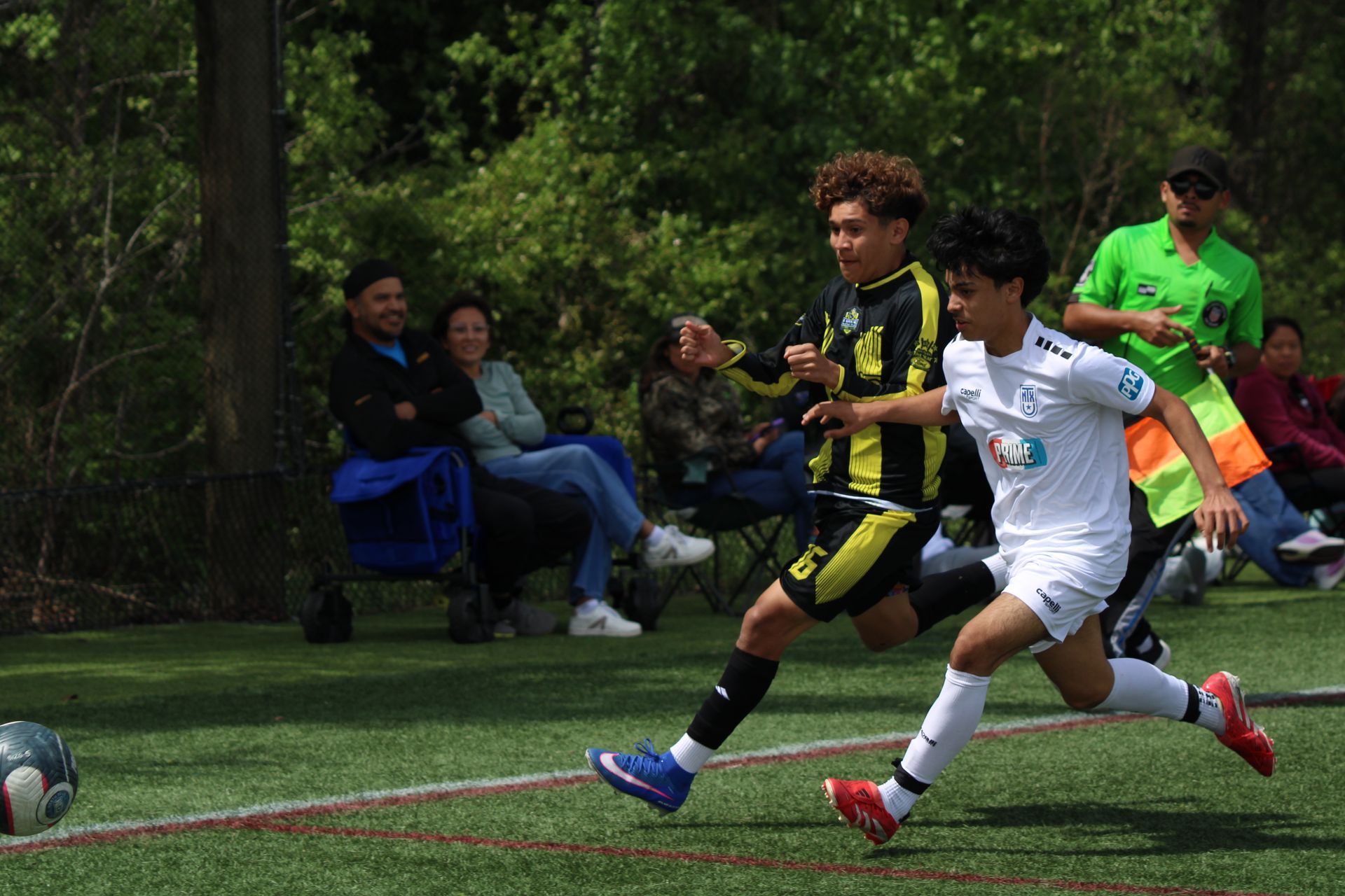Youth soccer players sprint after the ball on a grassy field, with spectators seated in the background.