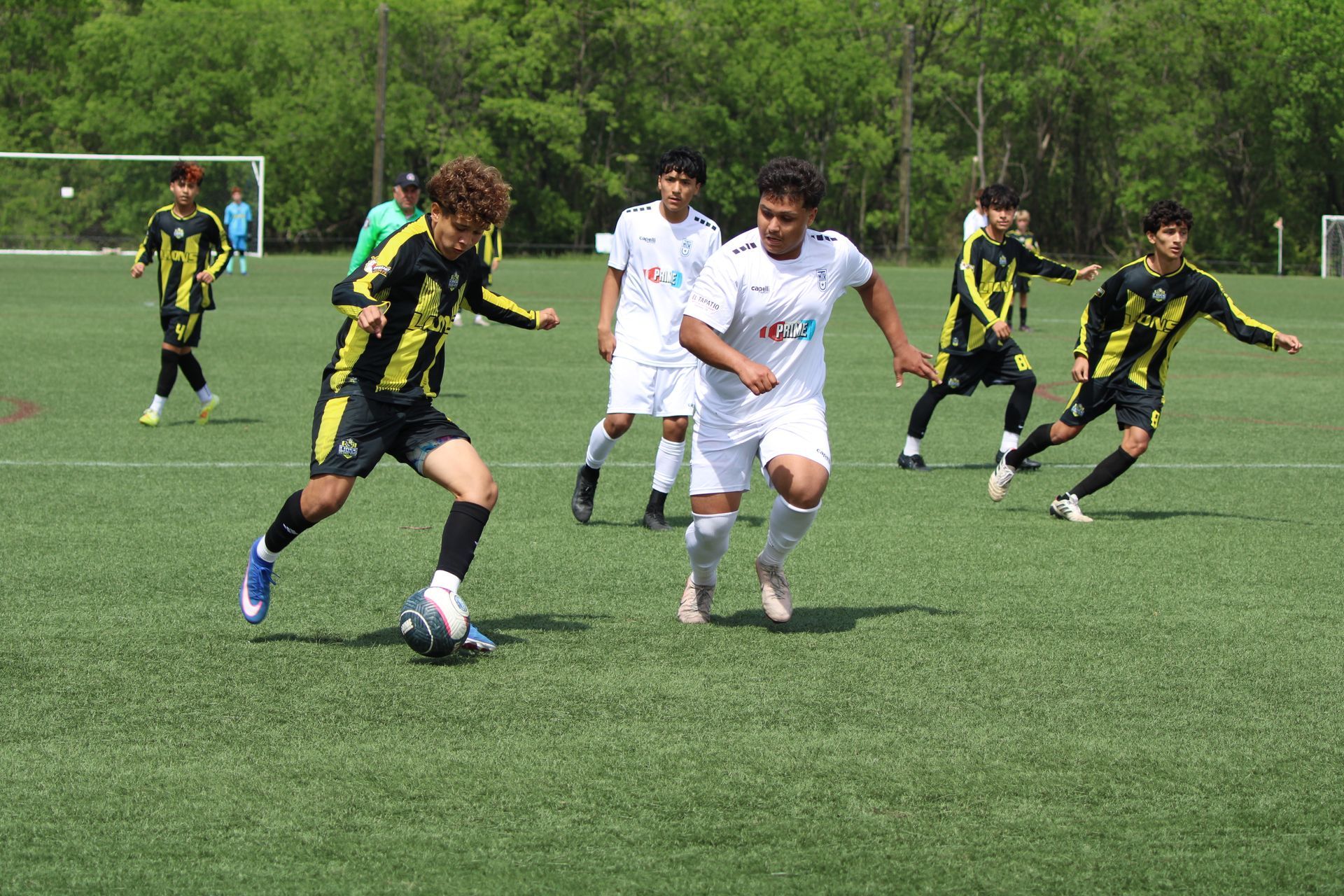 Soccer players in black and yellow and white uniforms sprint after the ball on a grassy field.