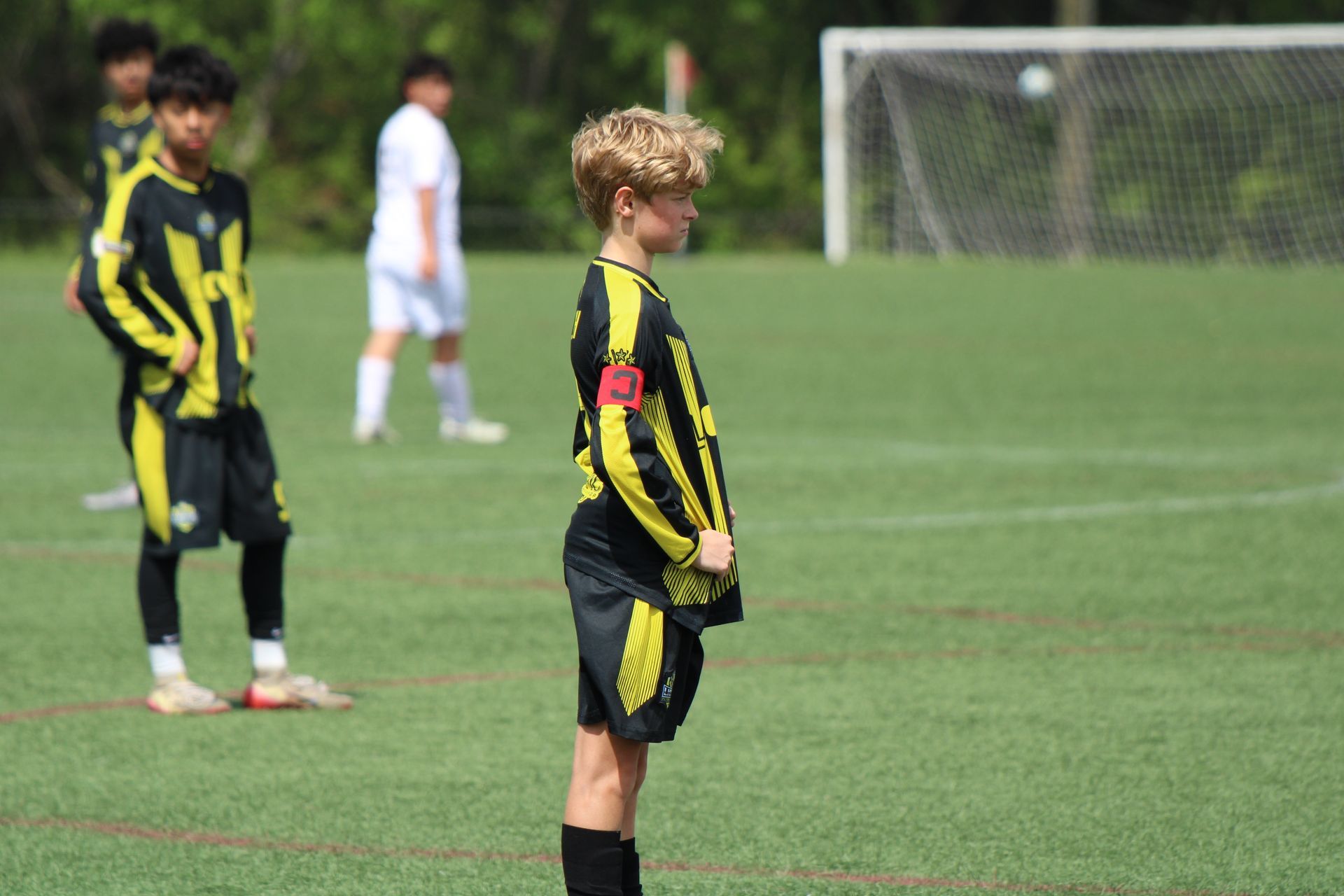 Youth soccer players in black and yellow uniforms standing on a grassy field, with a goal in the background