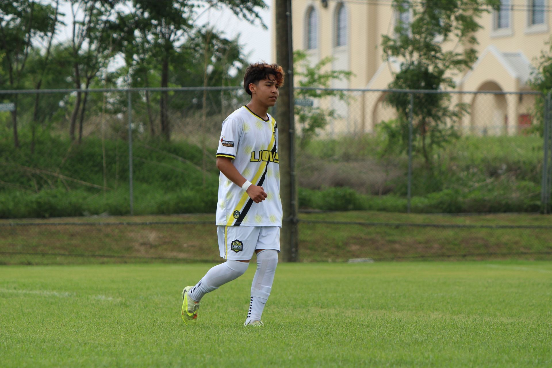 Soccer player in white uniform walking on a grassy field with trees and a building in the background