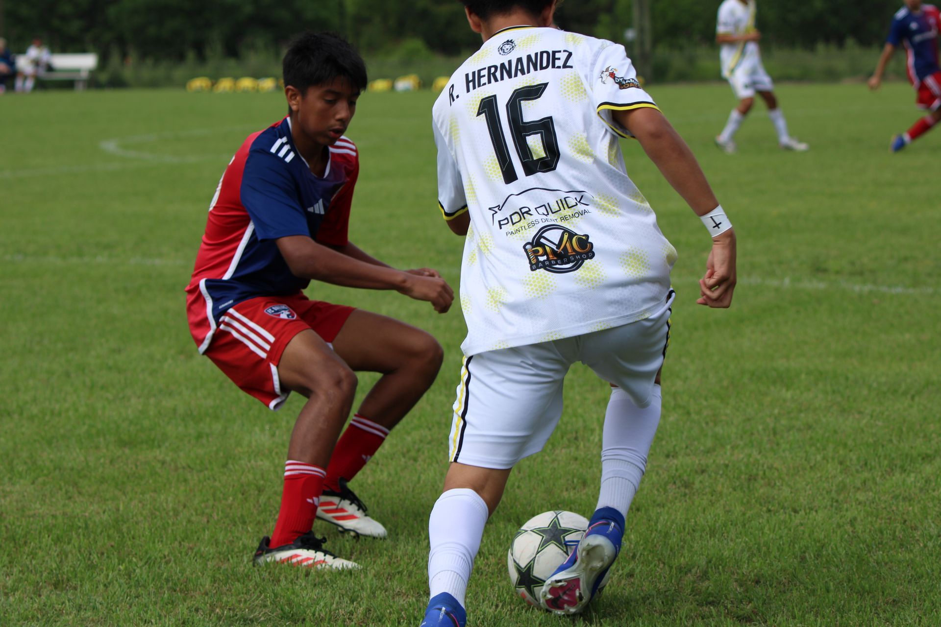 Two soccer players battle for the ball on a grassy field, one in red and blue, one in white.