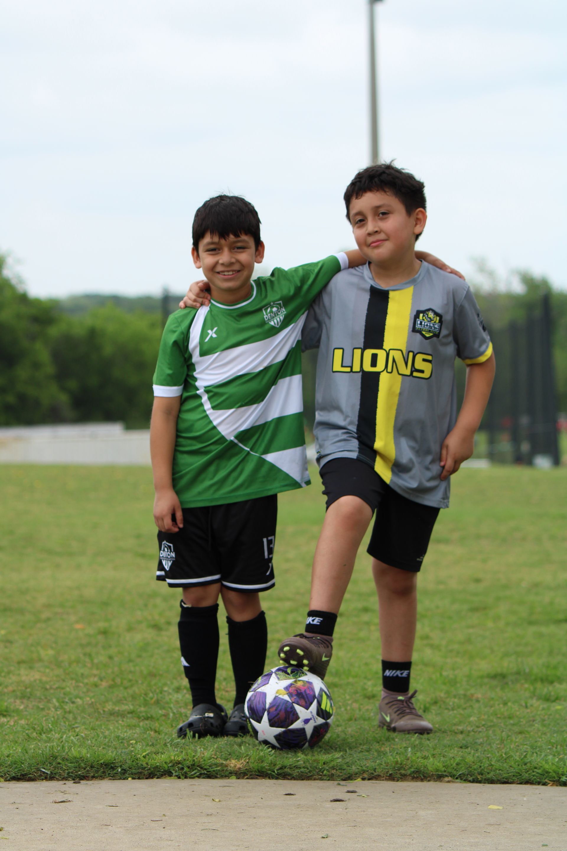 Two children in soccer jerseys standing with a soccer ball on a grassy field.