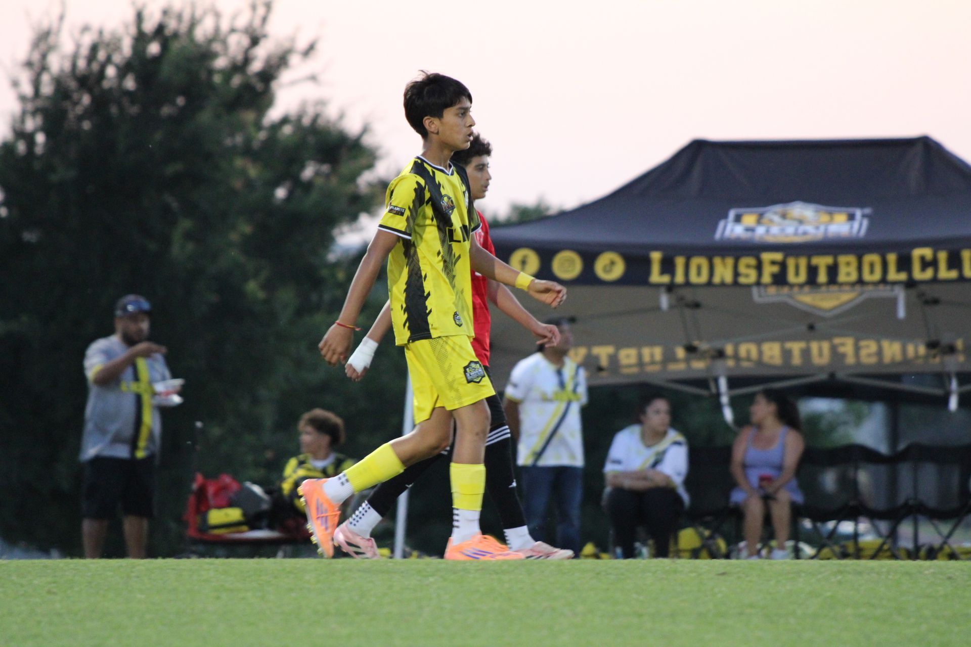 Soccer player in yellow and black uniform walking on field during a match, with tents and spectators in background
