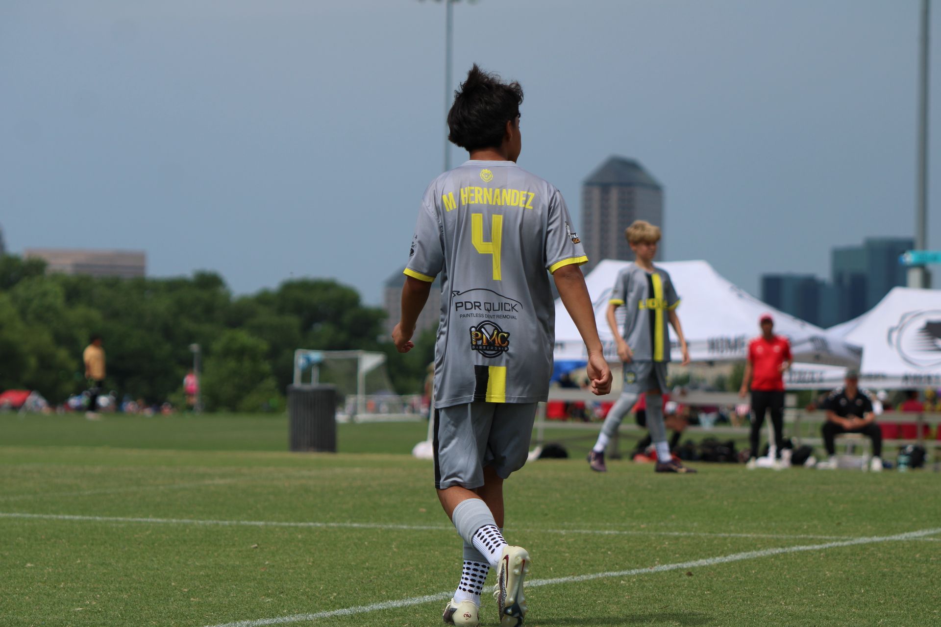 Soccer player in gray jersey with yellow number 4 walks on a grassy field near tents and other players