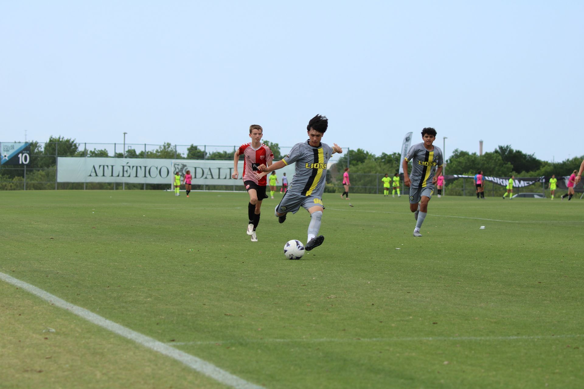 Soccer players in gray and yellow sprint after the ball on a grassy field near the sideline.