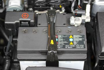 A black car battery under a hood, secured by a metal bracket, showing the positive terminal cap and warning labels.