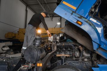 A mechanic in a black shirt and gray pants inspects the engine of a raised blue semi-truck inside a warehouse.