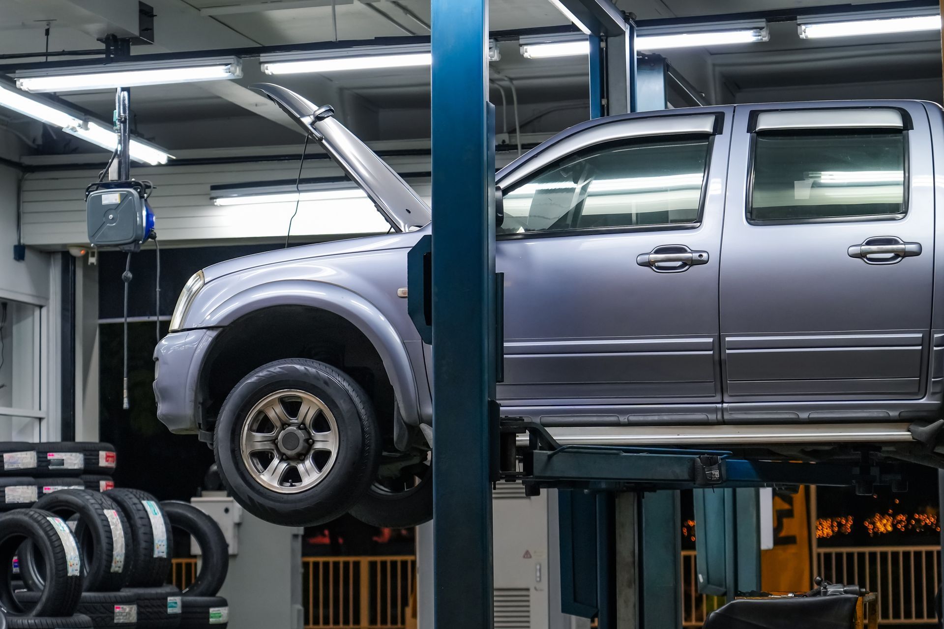 A gray pickup truck with its hood open is elevated on a lift inside an auto repair shop with tires visible in the foreground.