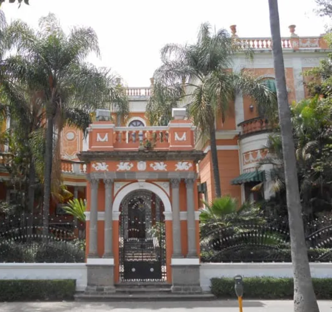Entrada a la mansión ornamentada en naranja y blanco con palmeras y una puerta negra.
