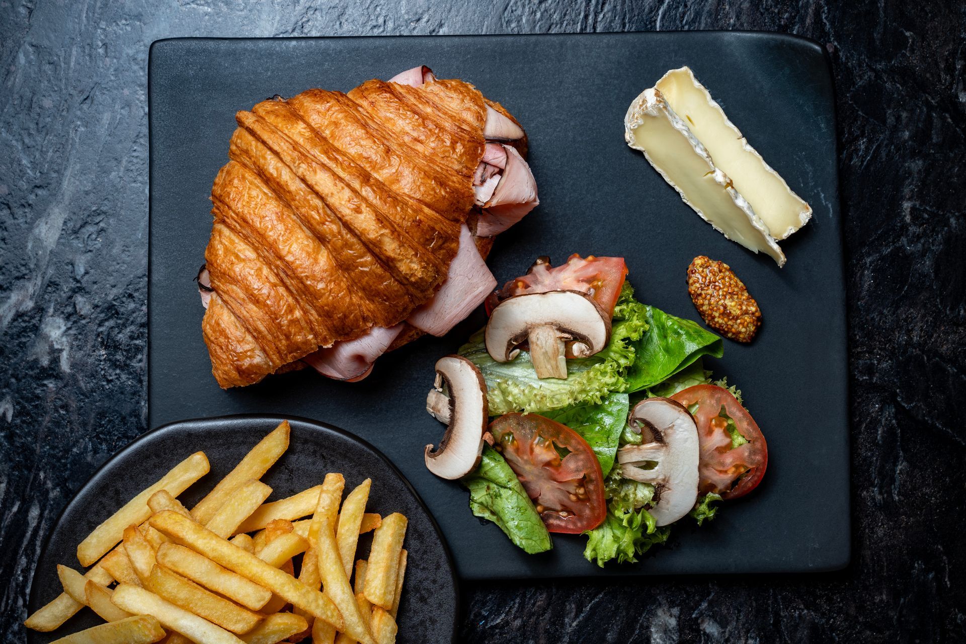 Sándwich de croissant, papas fritas, ensalada y mantequilla en una bandeja negra para una comida.
