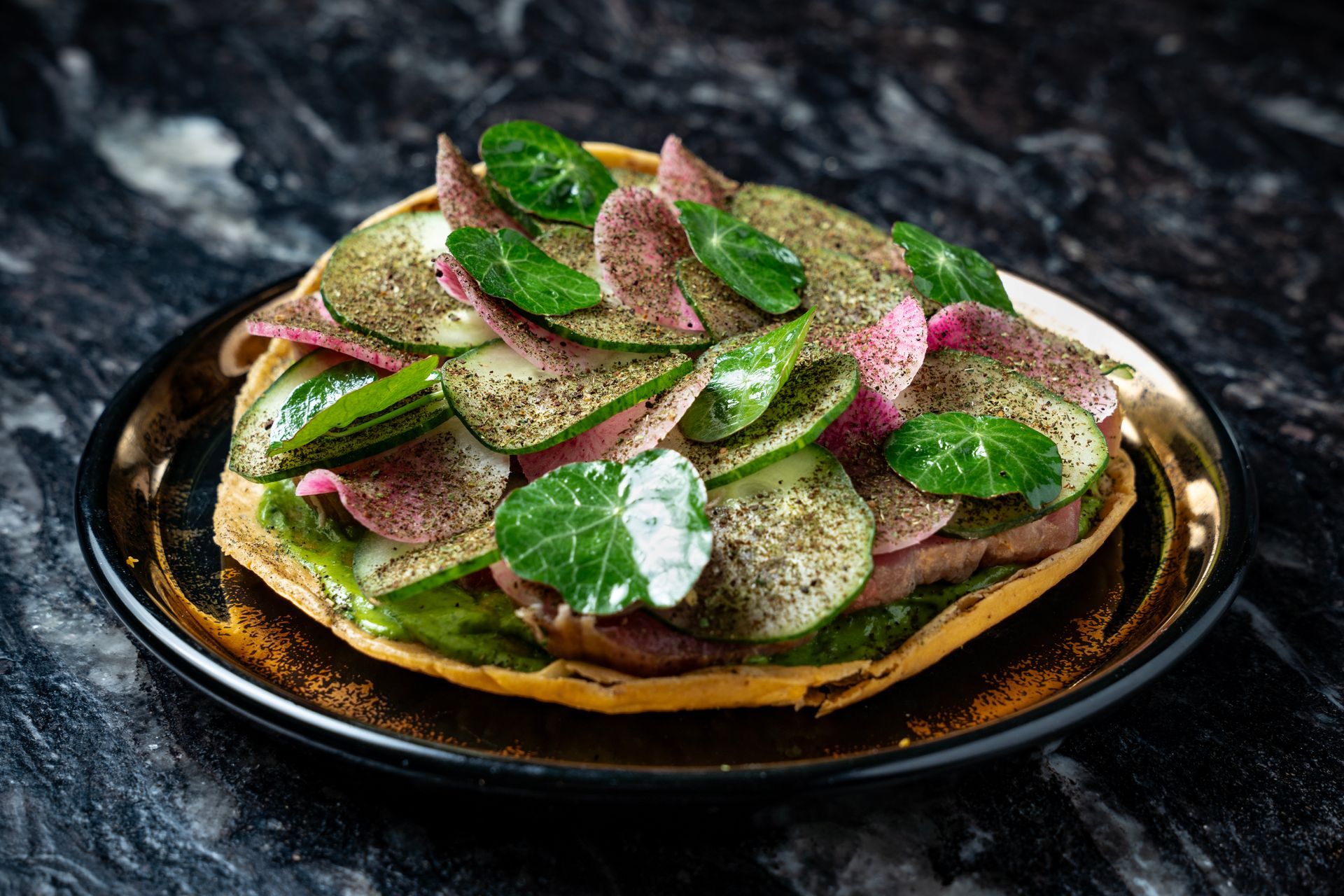 Pila de panqueques cubiertos con rodajas de manzana verde y crema rosa en un plato oscuro.