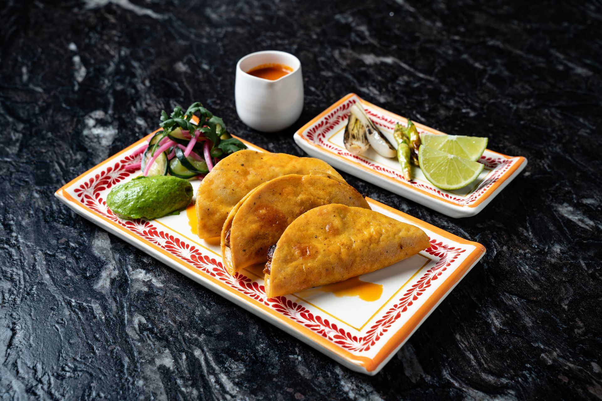 Tres quesadillas doradas en un plato decorativo con salsa verde, aguacate y salsa para mojar sobre una mesa oscura.