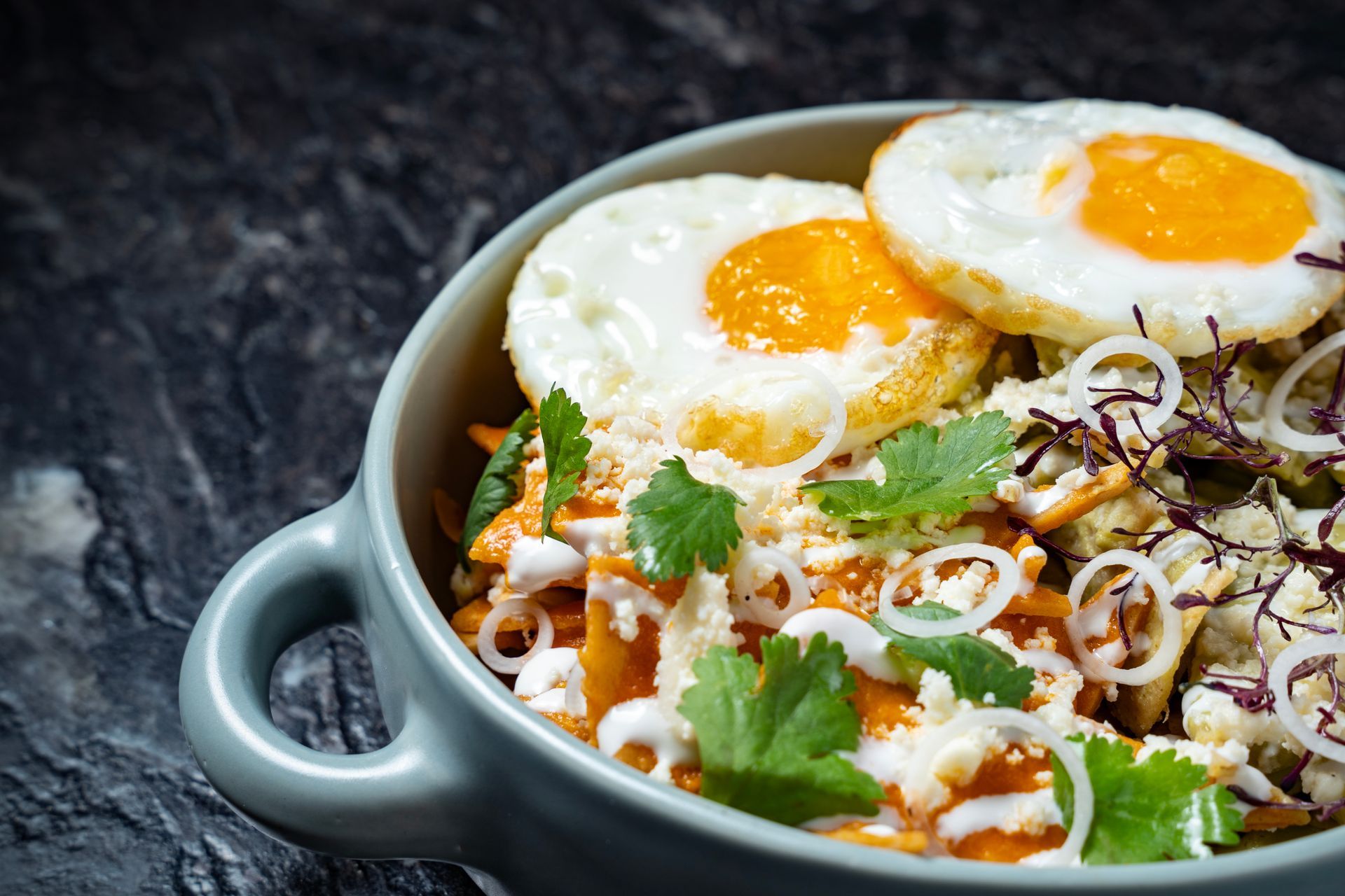 Un tazón de arroz cubierto con huevos fritos, hierbas y verduras ralladas sobre una mesa oscura.