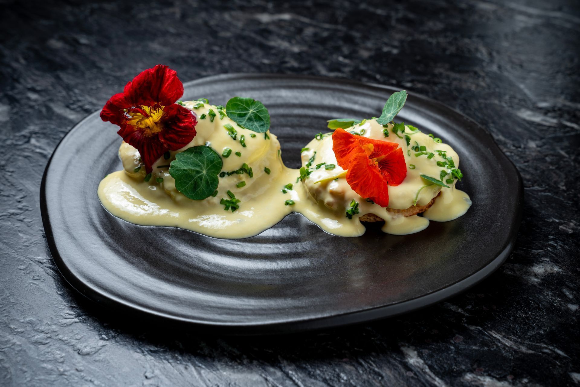 Dos aperitivos cremosos emplatados, adornados con hierbas y flores rojas sobre un plato negro.