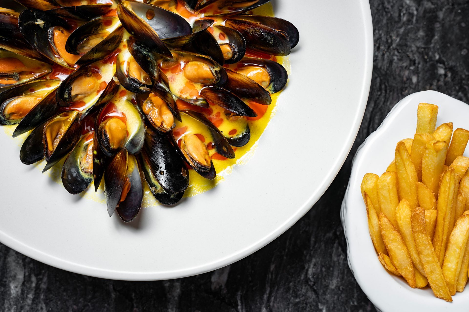 Mejillones en salsa servidos junto a un pequeño tazón de papas fritas en una mesa oscura.