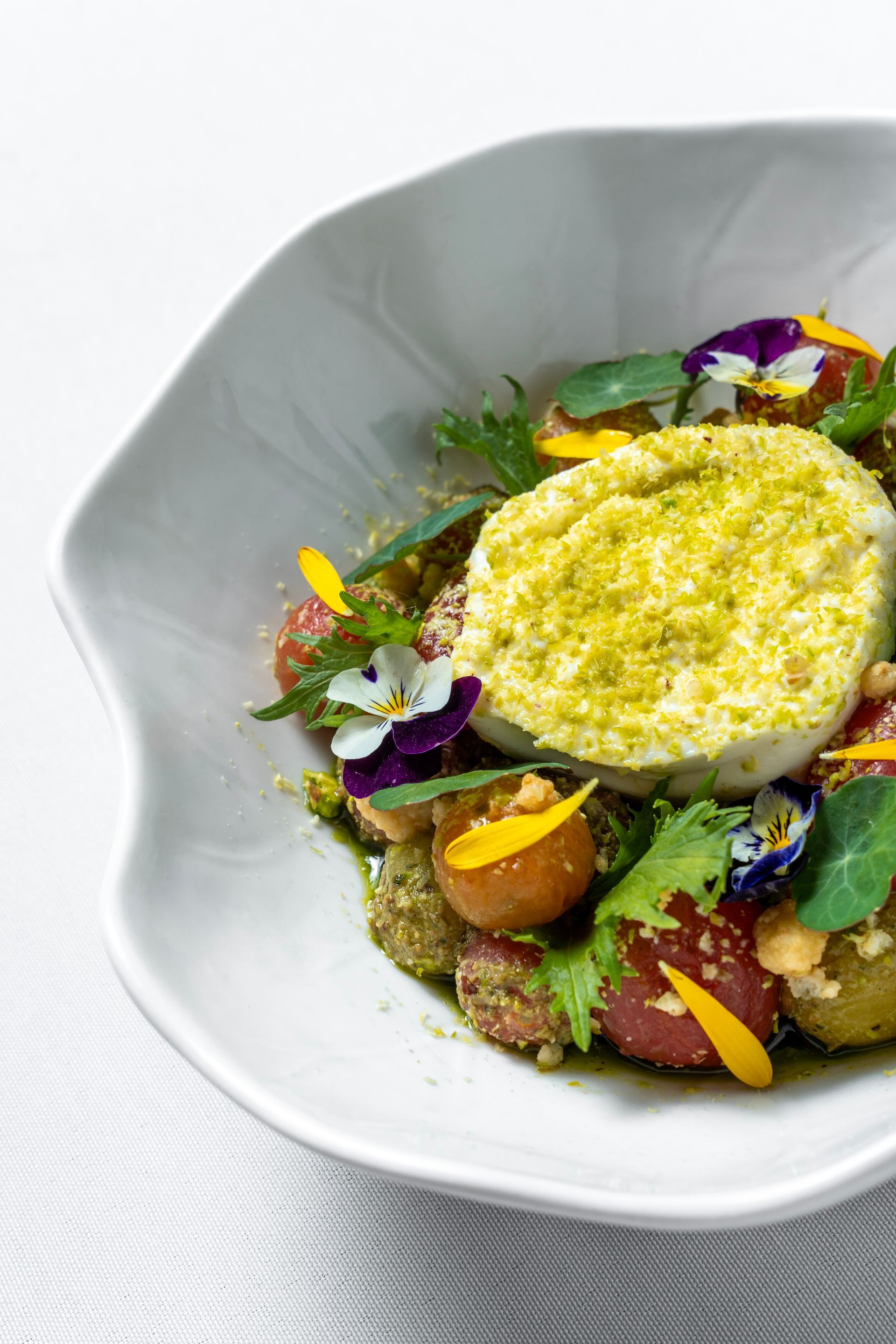 Plato gourmet con cobertura cremosa amarilla, verduras asadas y flores comestibles sobre un tazón blanco.