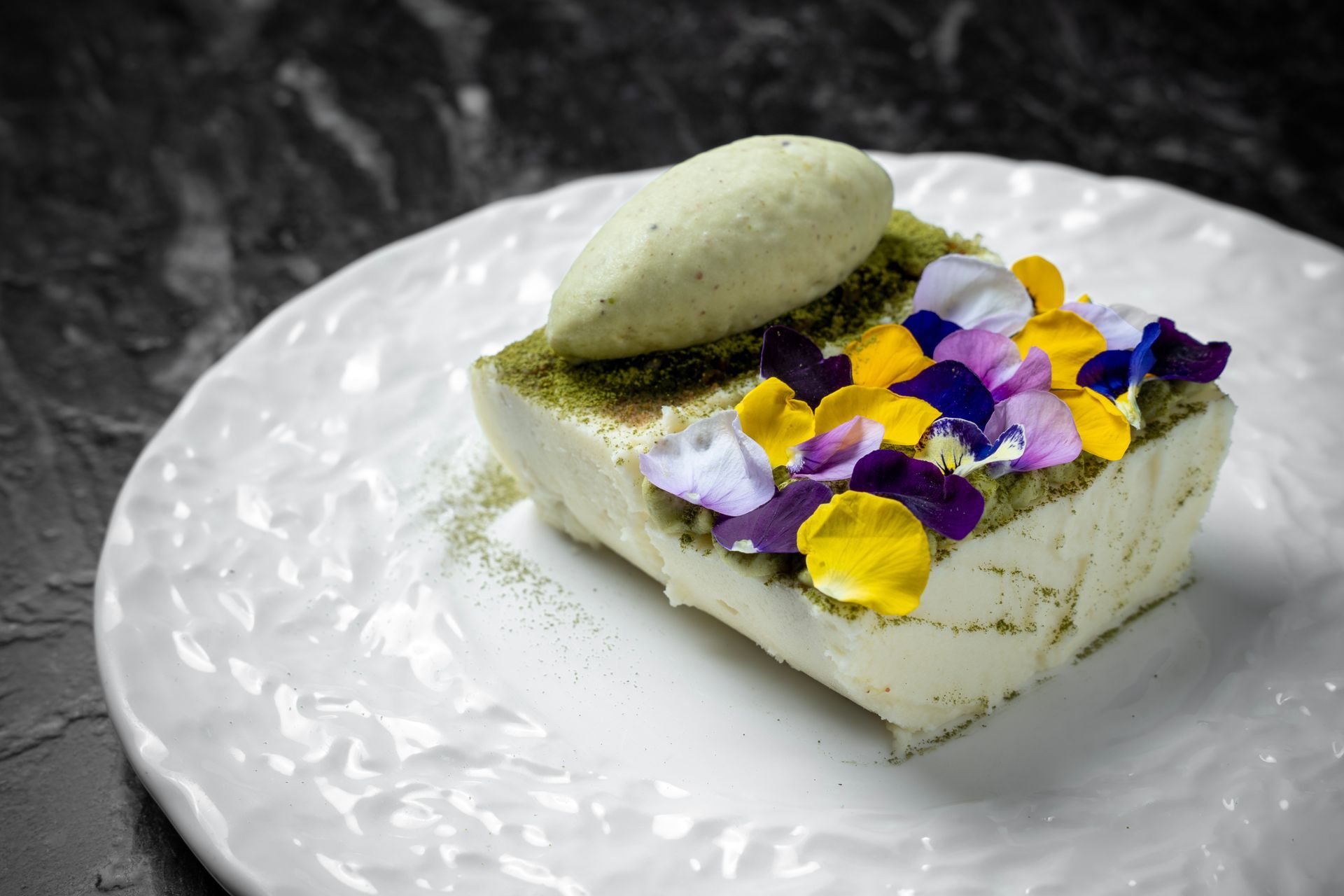 Postre cuadrado con mousse verde, decorado con flores comestibles moradas y amarillas sobre un plato blanco.
