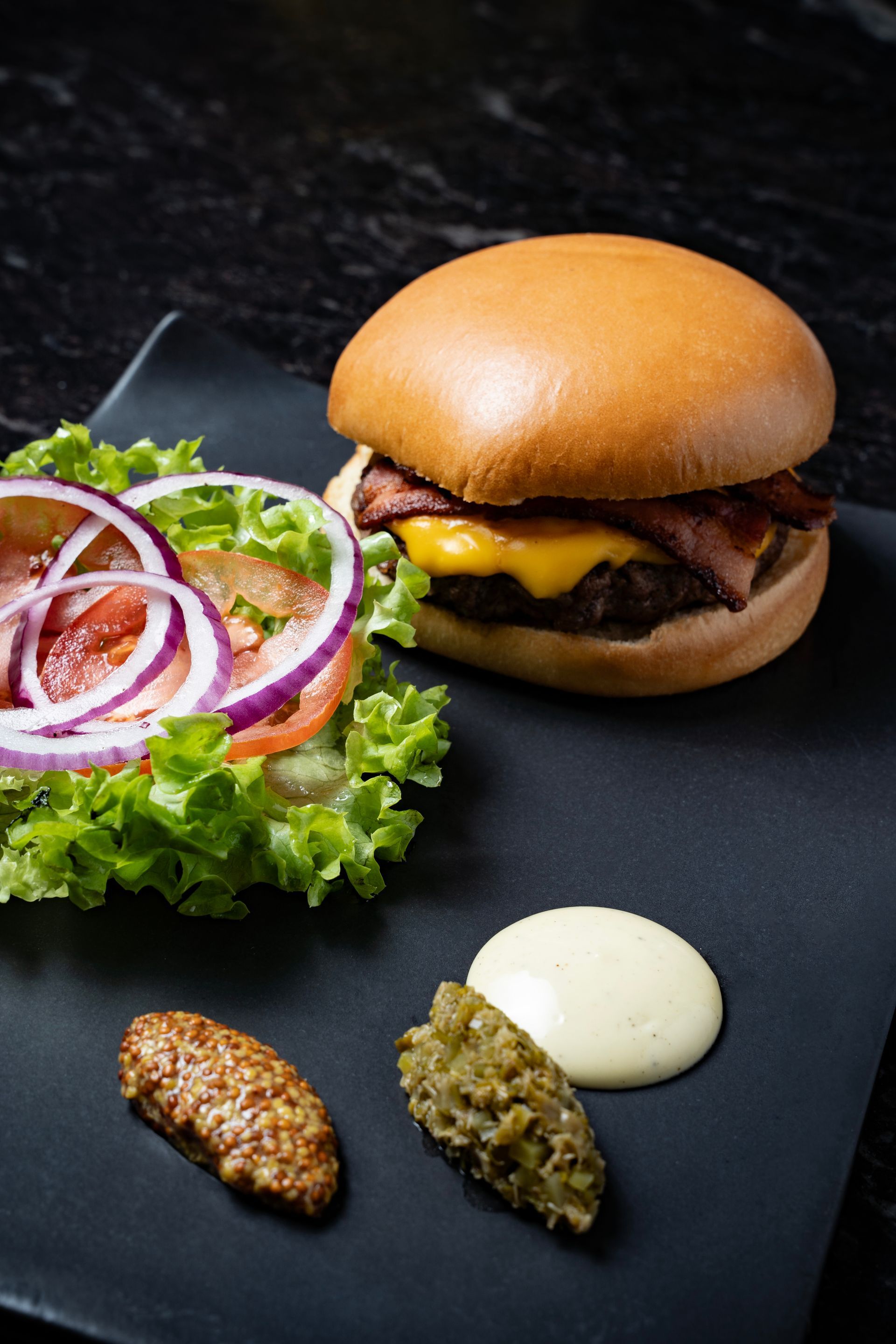 Hamburguesa con queso, lechuga y cebolla, además de salsas y chutney en un plato oscuro.