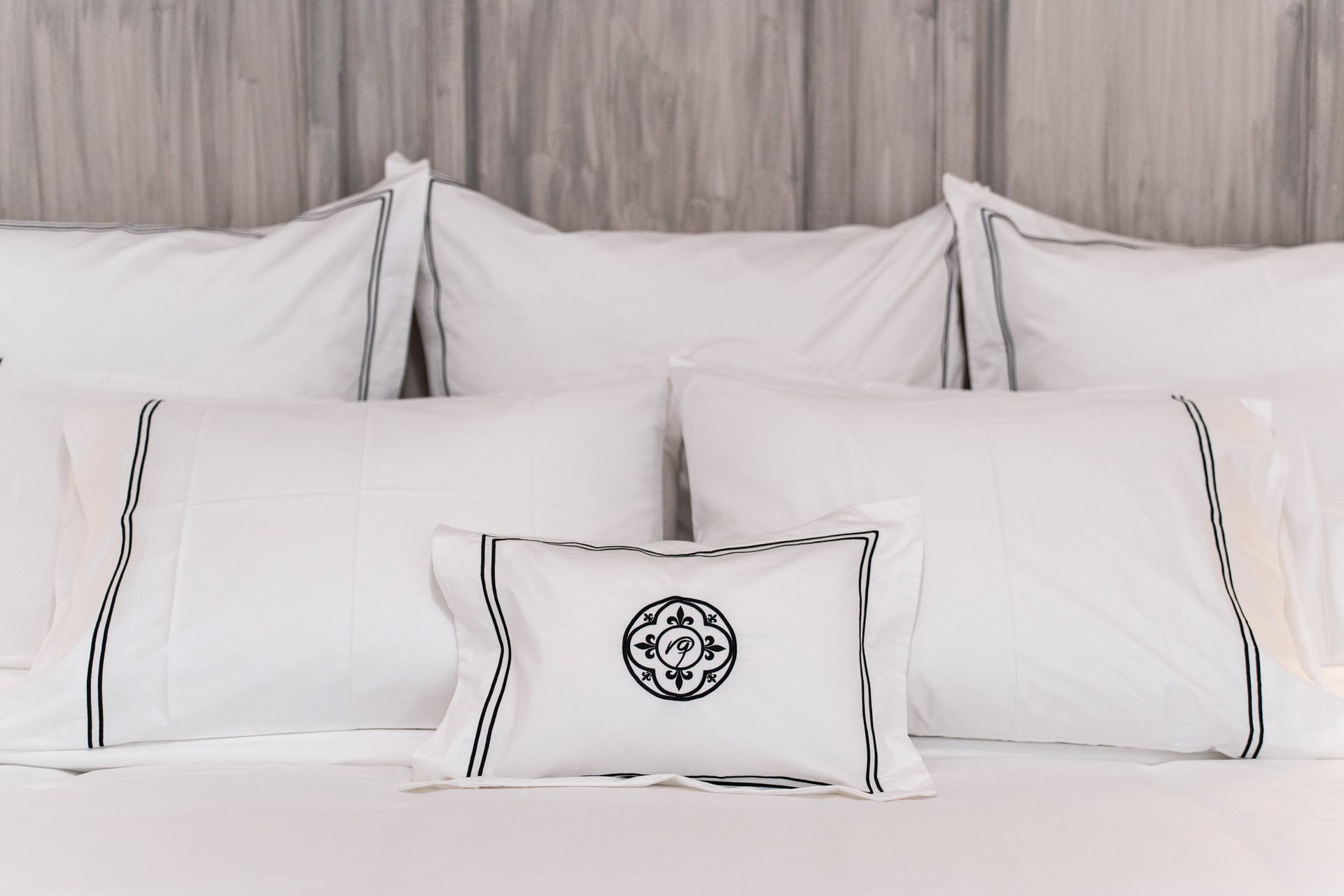 Cama blanca con almohadas cuidadosamente dispuestas y una almohada lumbar decorativa sobre un cabecero de madera clara.