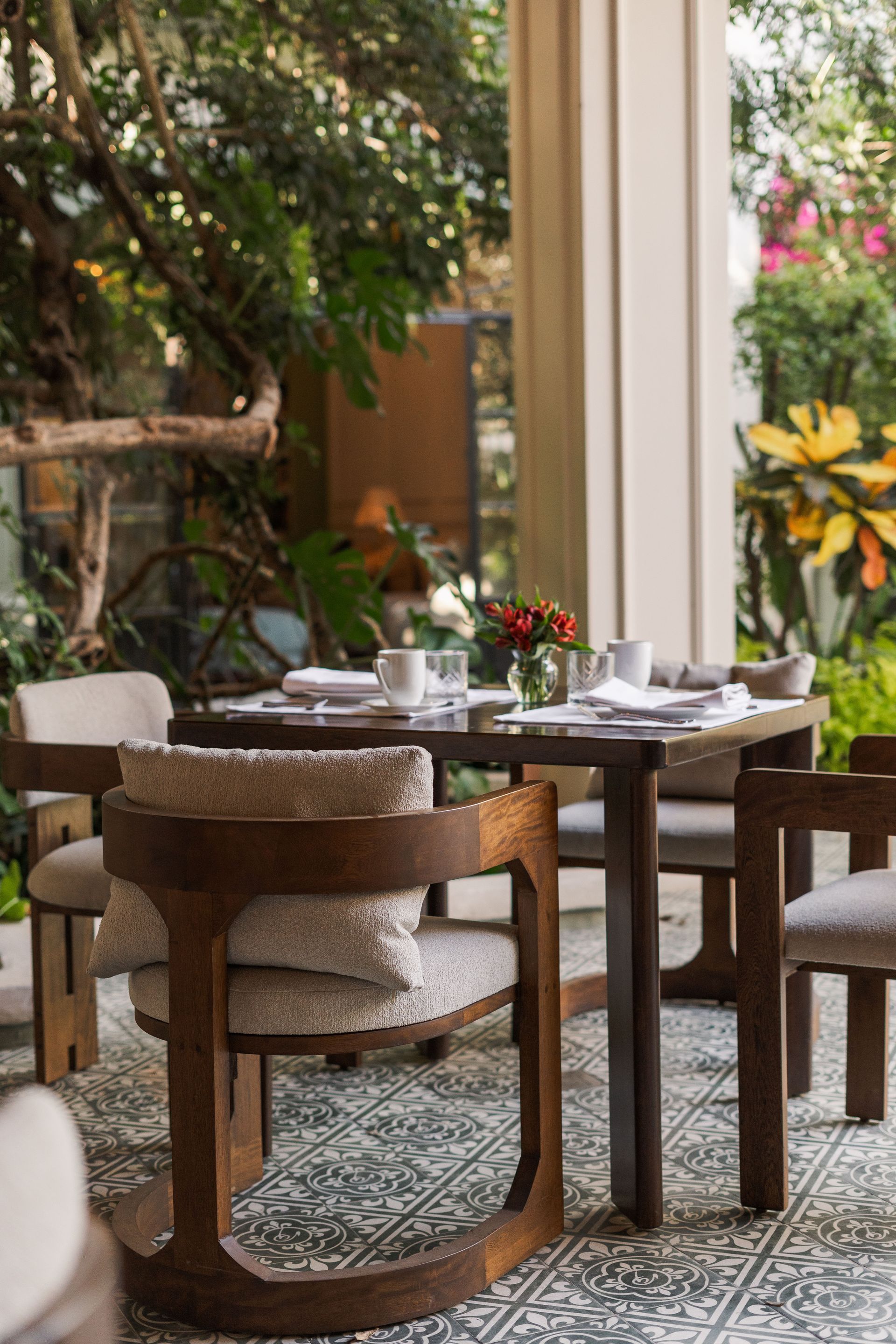 Mesa y sillas de café vacías junto a una ventana con vistas a un jardín exuberante.