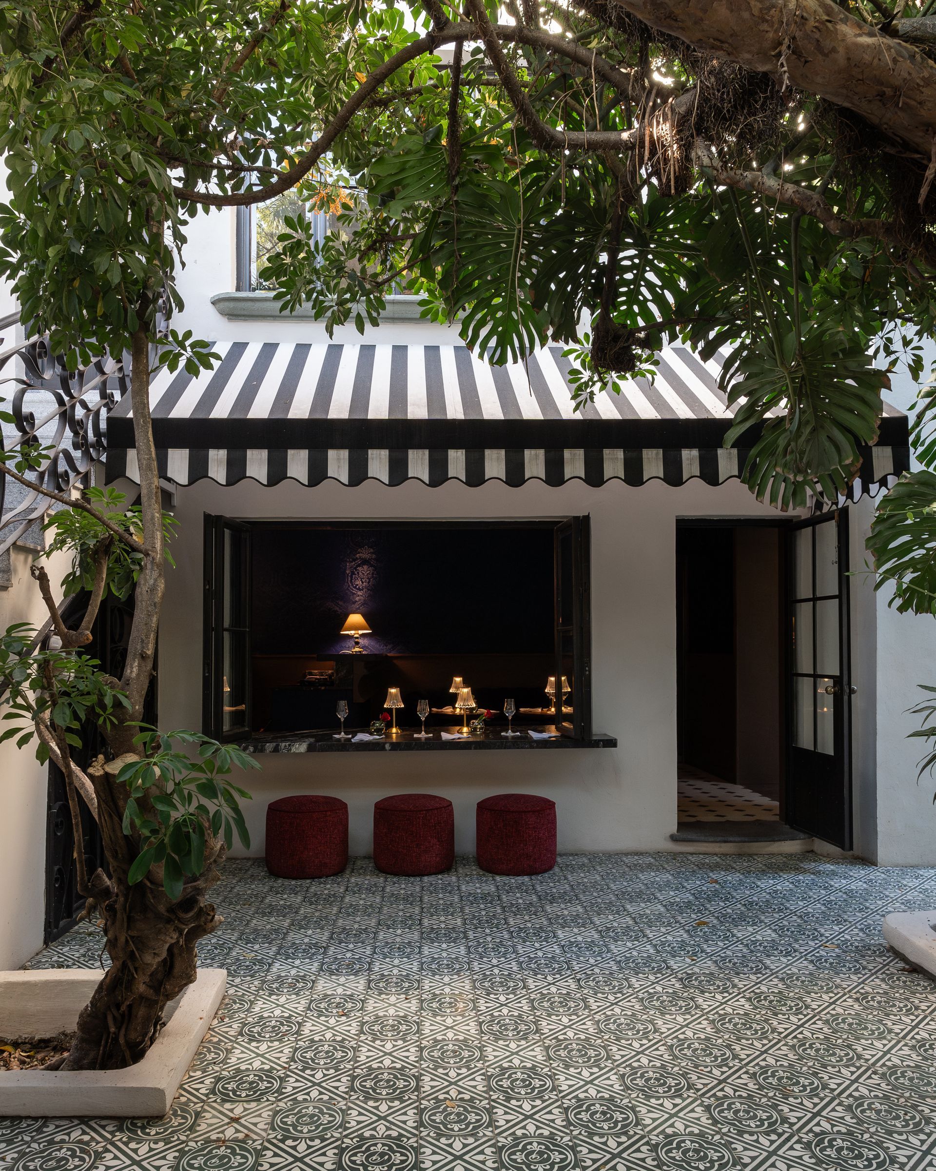 Fachada de cafetería al aire libre con toldo a rayas, ventana abierta, taburetes rojos y patio de baldosas estampadas bajo los árboles.