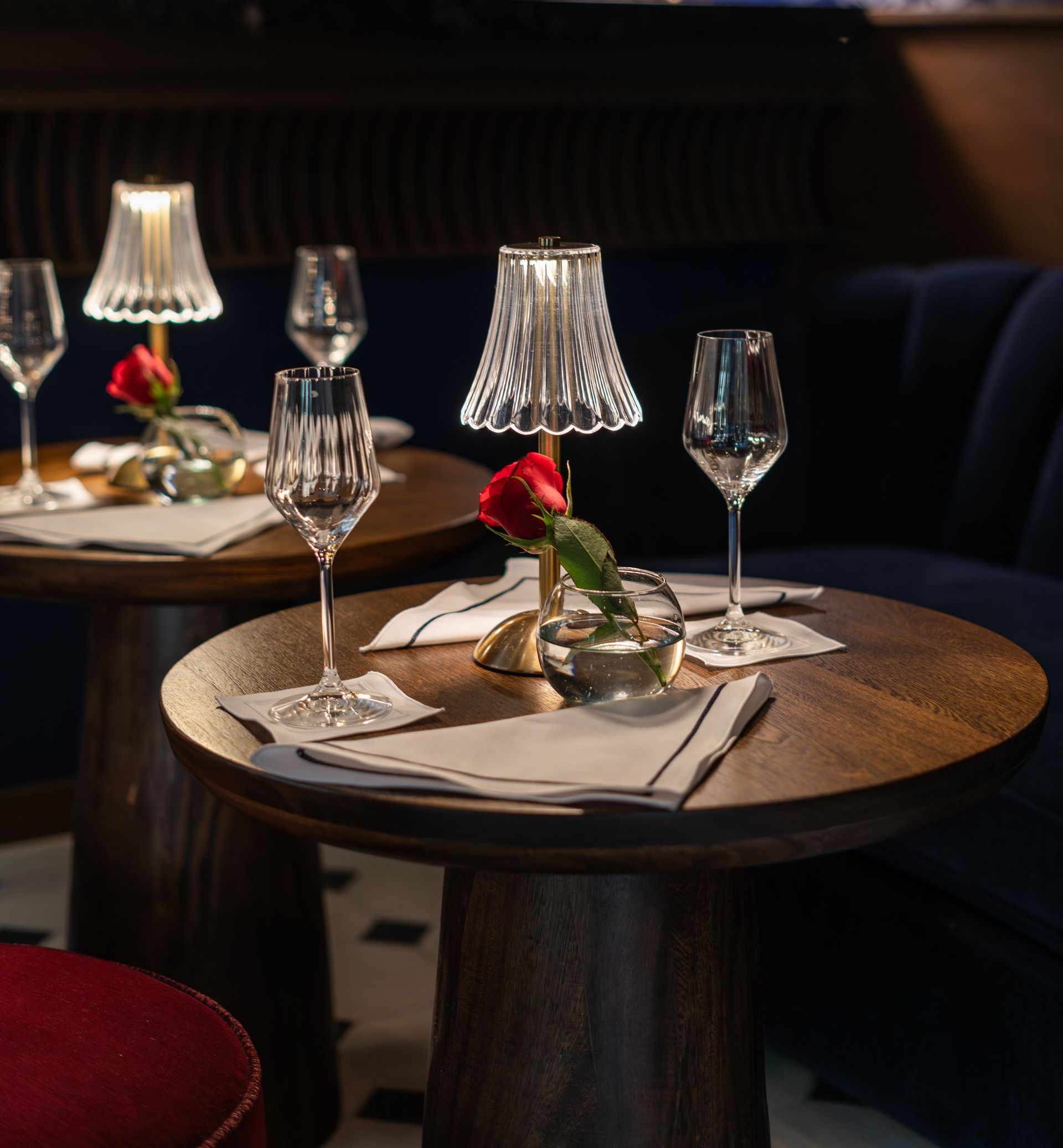Elegante mesa de restaurante puesta con copas de cristal, servilletas dobladas y centro de mesa de rosas rojas bajo una cálida luz de lámpara.
