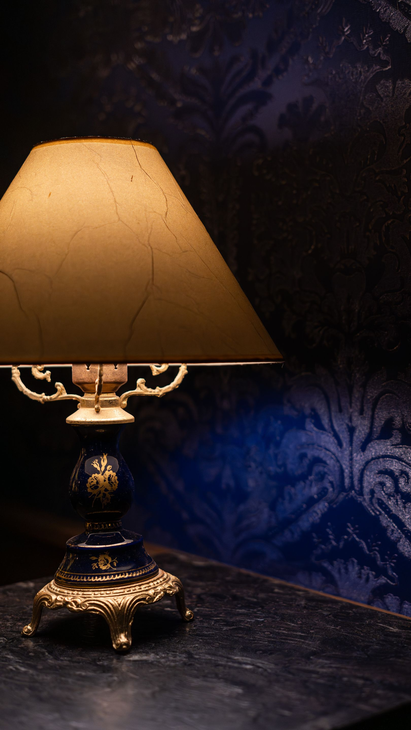 Lámpara de mesa ornamentada con pantalla beige sobre una mesa oscura frente a una pared estampada de color azul intenso.