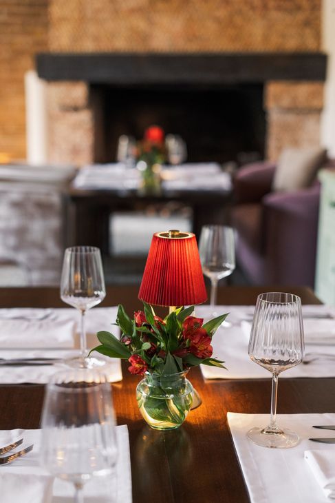 Elegante mesa de comedor con lámpara roja como centro de mesa, copas de vino y chimenea al fondo.