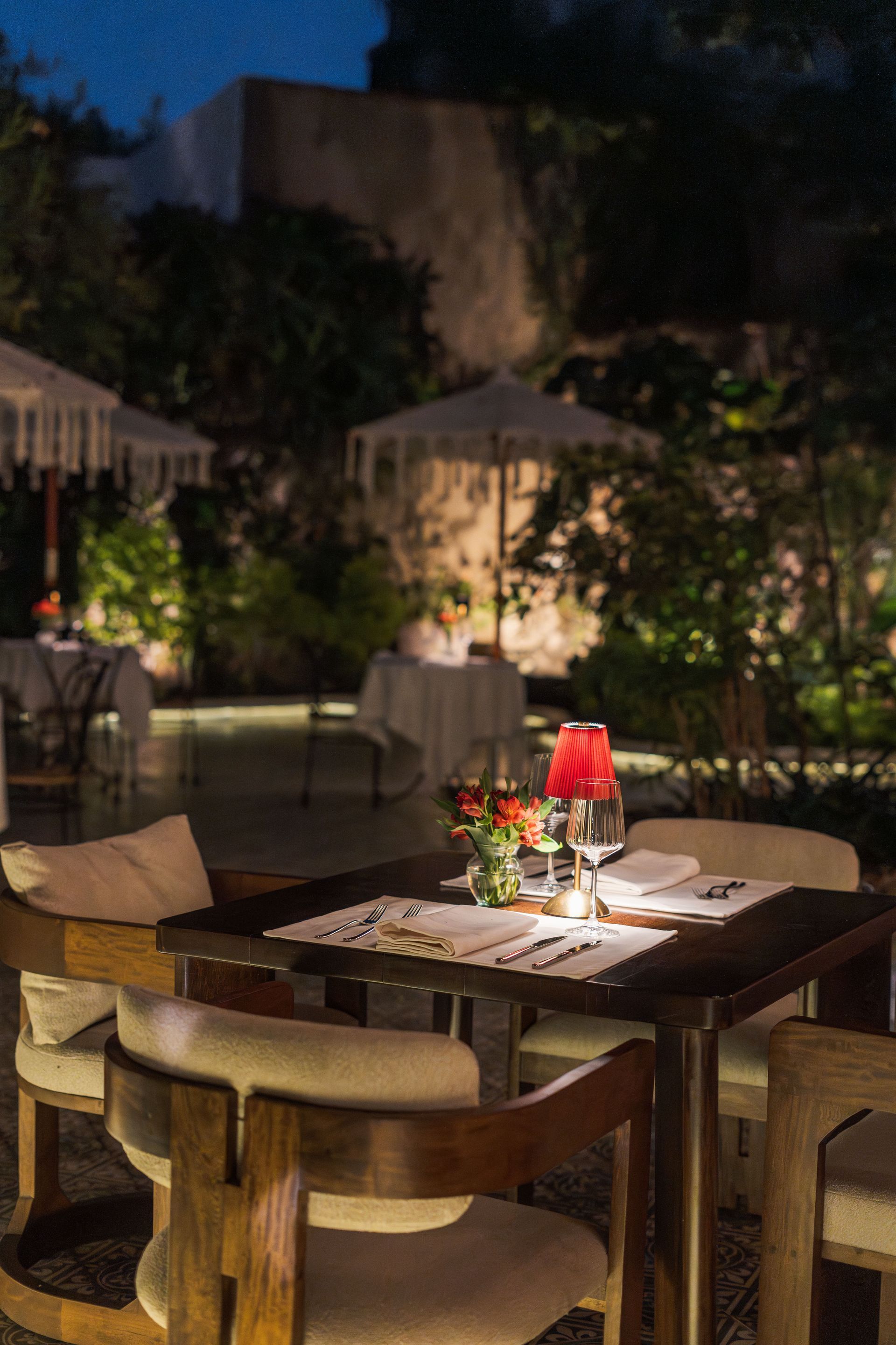 Mesa de comedor en un patio exterior iluminada por una lámpara roja por la noche, con sillas y plantas al fondo.